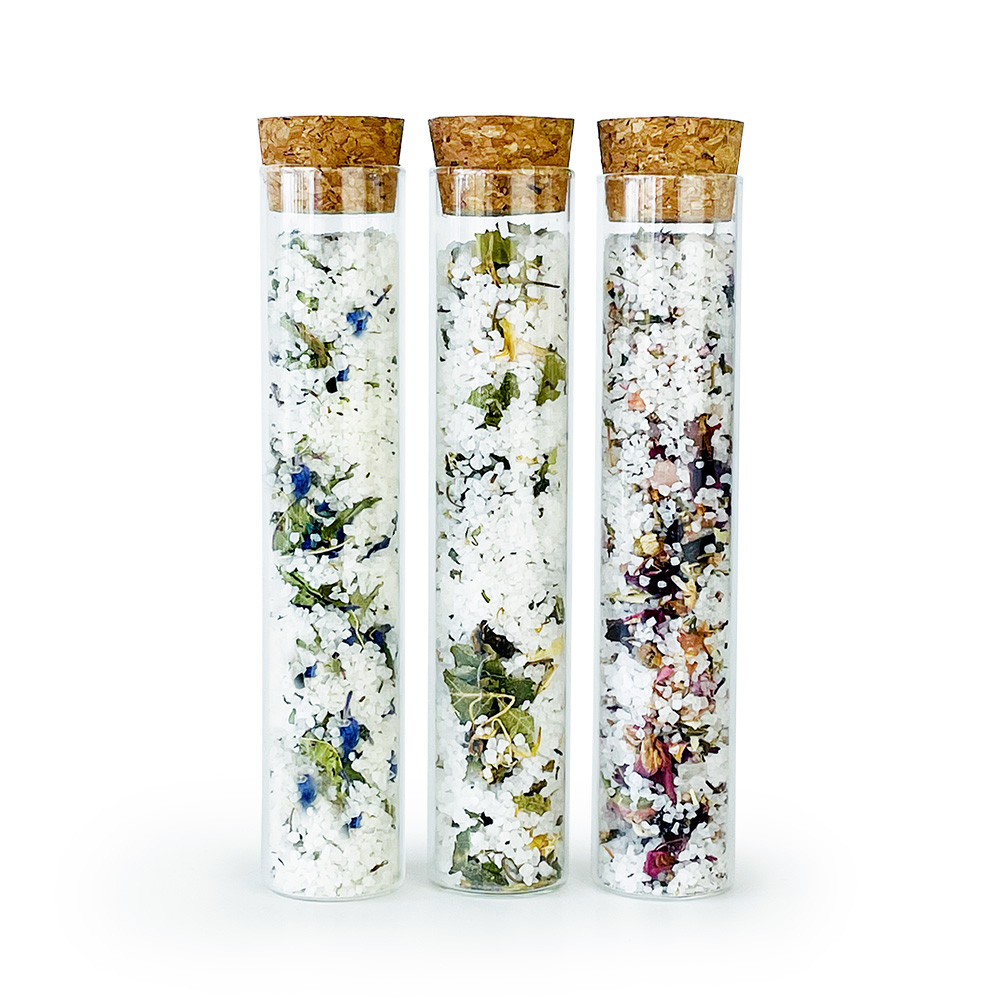 SP-AB-1_10101392096_02_LOW Drei Glasröhrchen mit Korkverschluss, gefüllt mit verschiedenen Badesalzen und getrockneten Blumen.