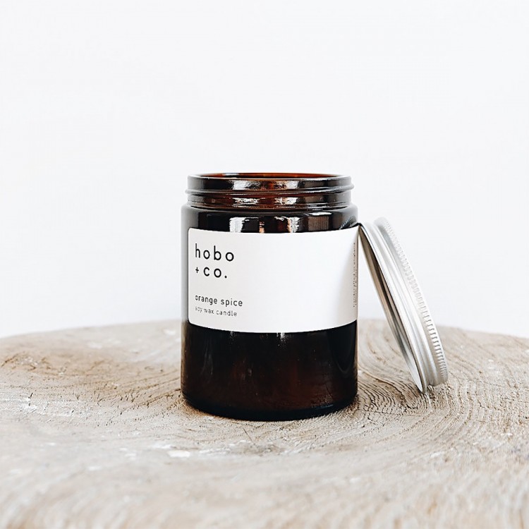 IMG_6553 Braunes Glas mit weißem Etikett und silbernem Deckel, enthält eine Duftkerze von hobo + co. auf einem Holzuntergrund.