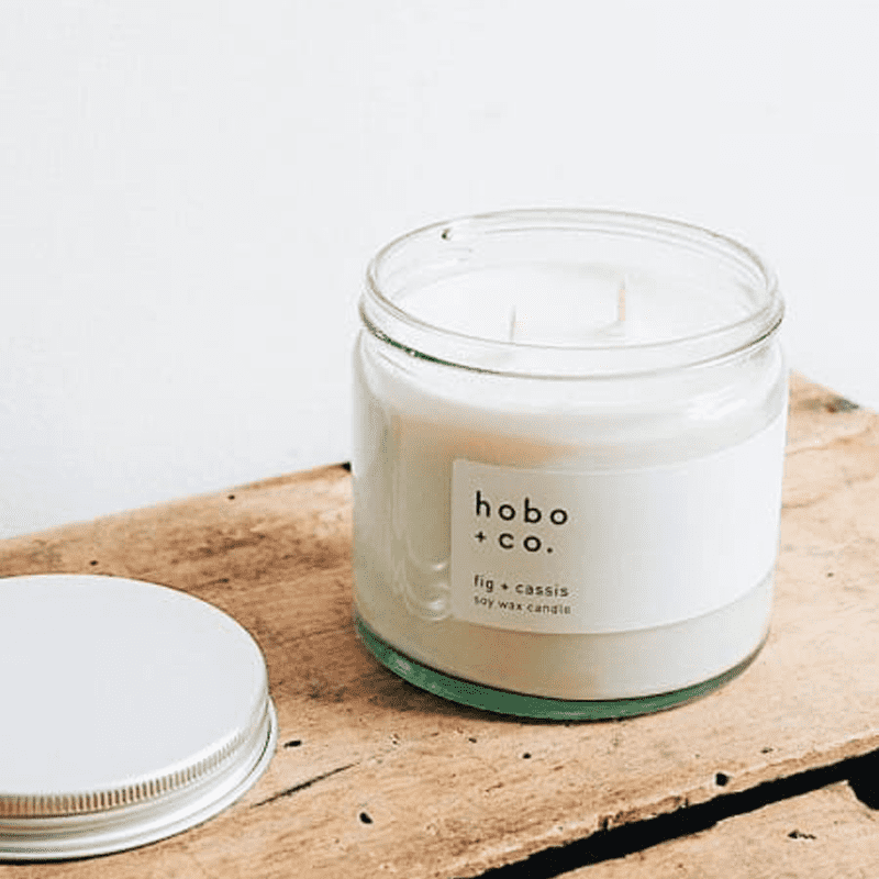 hobo-co-fig-cassis-essential-oil-candle-250ml Hobo + Co. Fig & Cassis Duftkerze in einem Glasbehälter auf einem Holztisch, Deckel daneben.