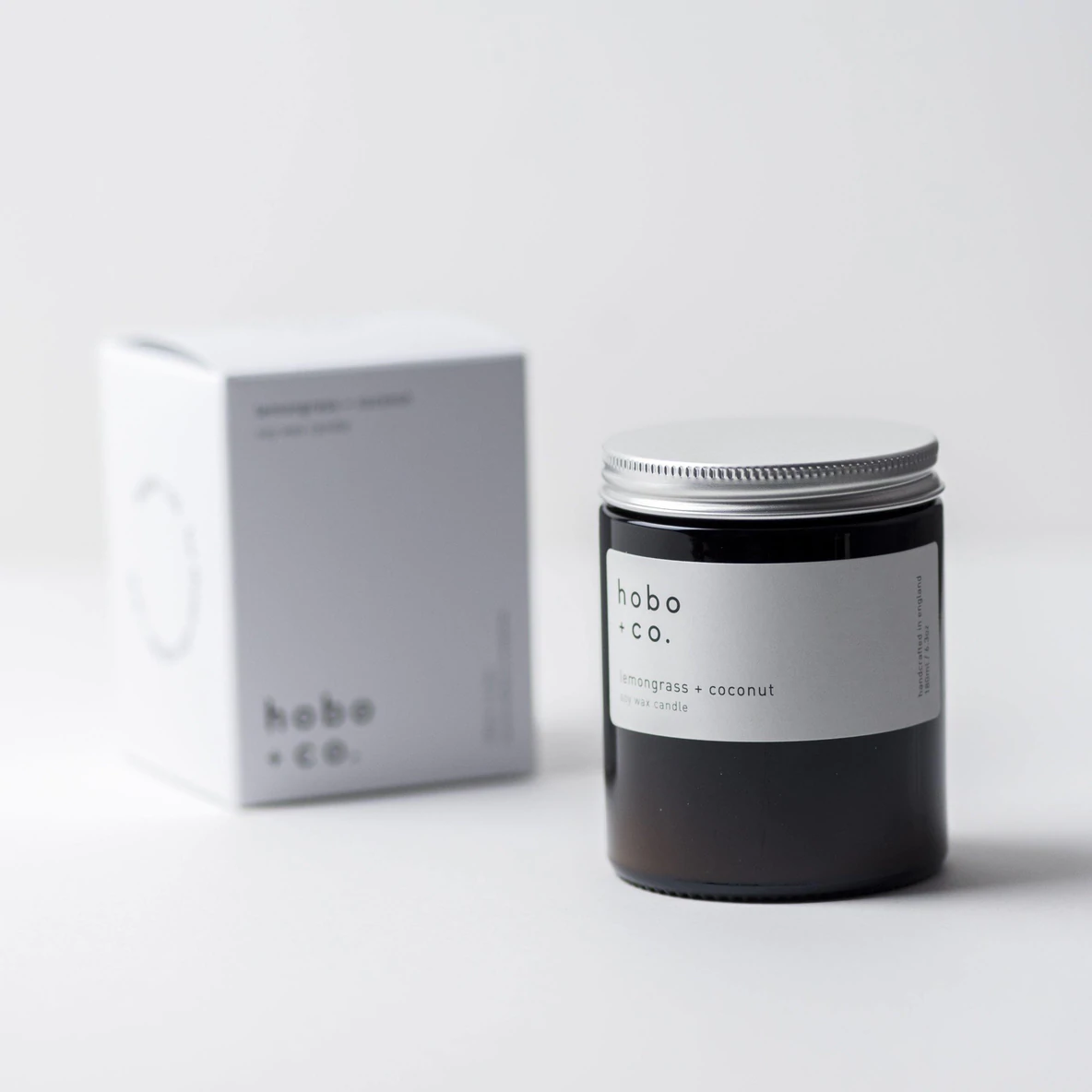3CBD172F-6A3D-4163-93A9-4457F8B30709 Eine Duftkerze von hobo + co. mit Zitronengras und Kokosnuss, neben der Verpackung auf einem weißen Hintergrund.