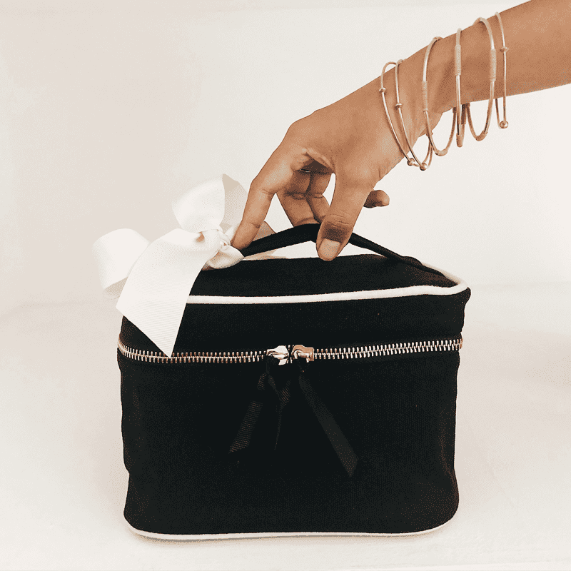 bag-all-beauty-box-klein-ohne-schrift-schwarz_1_optimizedsKSBmv8Vt3HXV Hand greift nach schwarzer Kosmetiktasche mit weißer Schleife auf weißem Hintergrund.