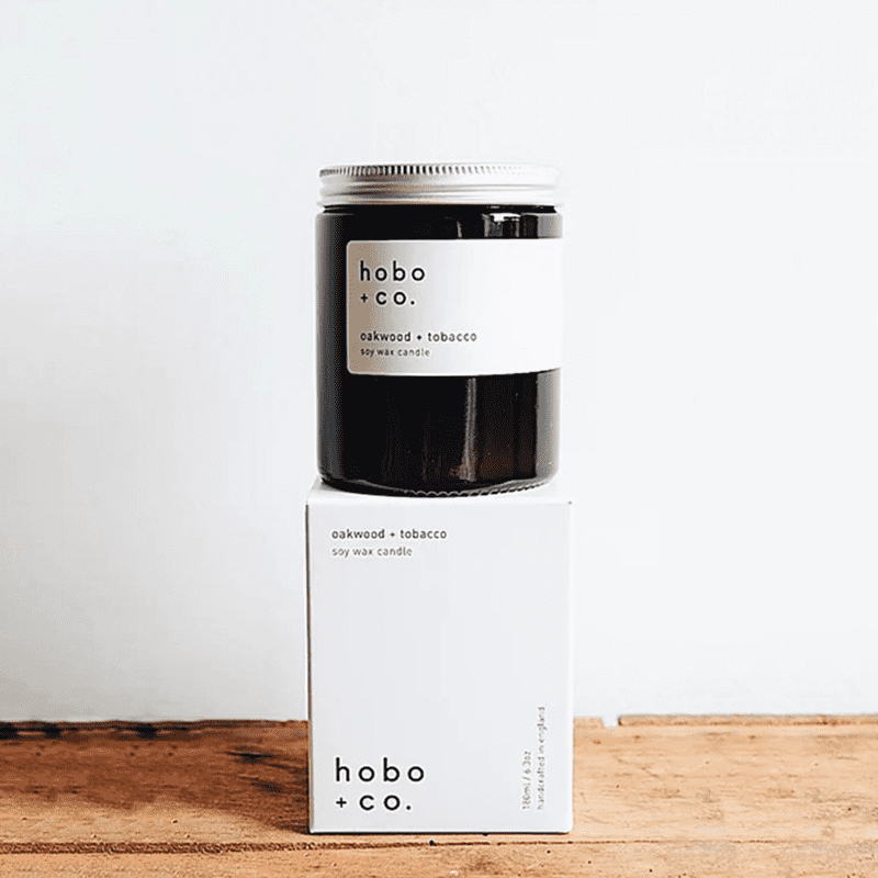 hobo-co-oakwood-tobacco-essential-oil-candle-180ml_optimized Hobo + Co. Oakwood & Tobacco Duftkerze auf Holzoberfläche, 180ml Glas und Verpackung.