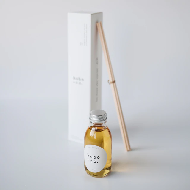 B2FA7882-E2A9-401D-9A9C-2EF7572F1F83 Kleine Flasche mit Duftöl von hobo + co. und Holzstäbchen vor einer weißen Verpackung auf einem weißen Hintergrund.